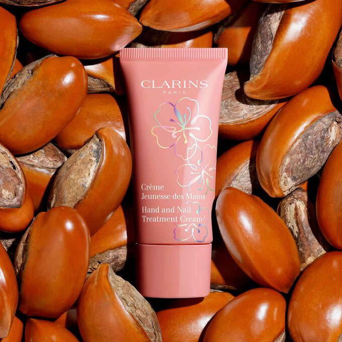 Plan produit de la Crème Mains Floraison Féerique Clarins entourée de graines de beurre de karité, mettant en valeur sa formule à 90 % d'origine naturelle.