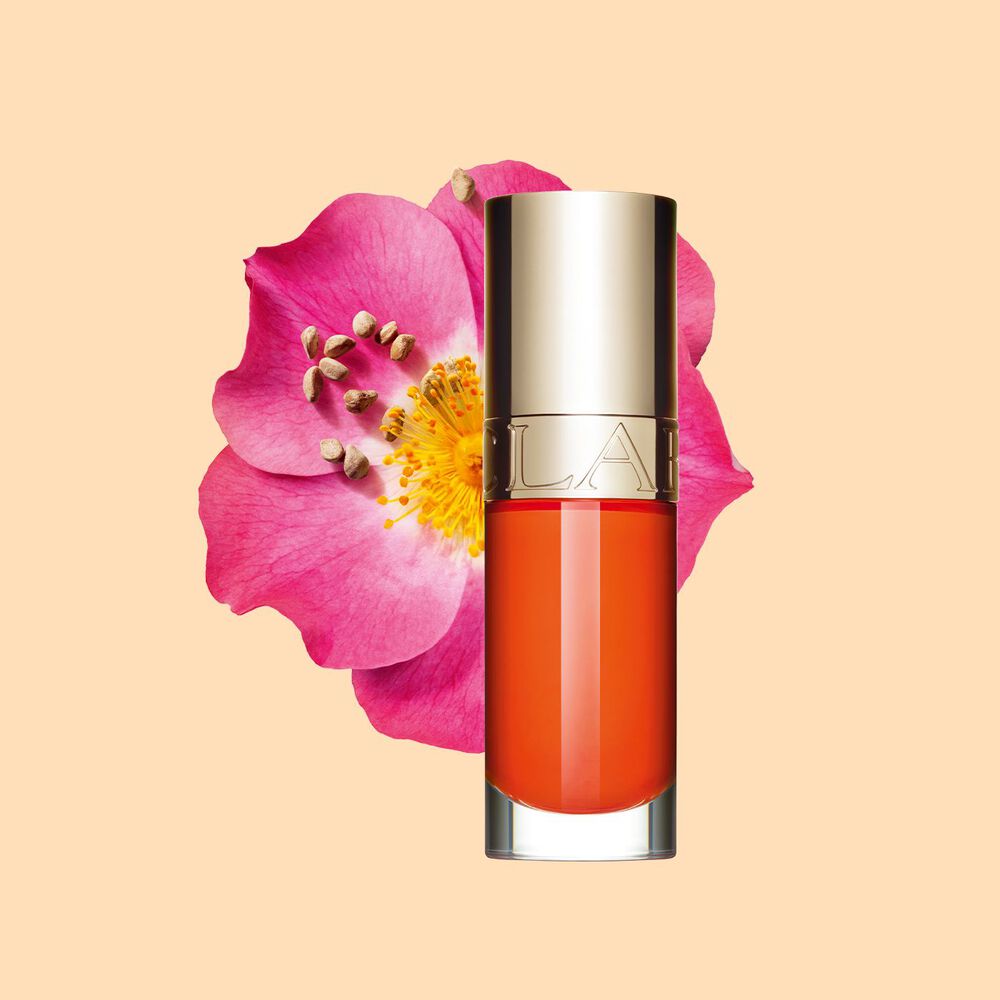 Plan produit de l'huile lèvres orange foncé Clarins devant une fleur de rosier rouillé sur fond blanc
