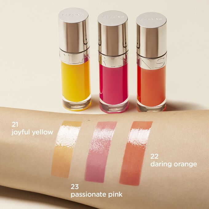 Présentation de la gamme huile lèvres Clarins jaune, rose et orange. Un échantillon de chacune a été apposé sur le bras d'une femme pour montrer le rendu du produit